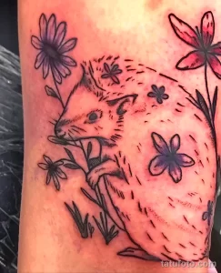 Фото тату ежика, окруженного ромашками и другими цветами. Рисунок выполнен в черно-белой гамме с использованием штриховки.