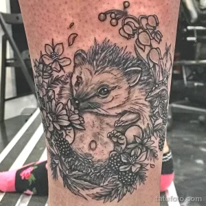 Фото детализированной татуировки с изображением ежика, окруженного цветами и бабочками. Черно-белый рисунок на ноге.