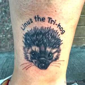 Фото татуировки ежика с надписью "Linus the Tri-hog". Милый дизайн, черные чернила, игривый шрифт. Идеально для любителей ежей и персонализированных тату.