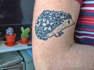Фото тату ежика, окруженного цветочной рамкой на руке. Черно-белый рисунок с детальной проработкой.