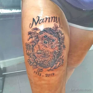 Фото тату ежика в окружении цветов с надписью "Nanny" и датами. Персонализированный рисунок татуировки в память о близком человеке.