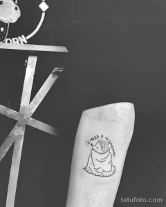 Фото тату маленького ежика с надписью "I need a hedgehug". Простой черно-белый рисунок в минималистичном стиле. Расположен на руке.