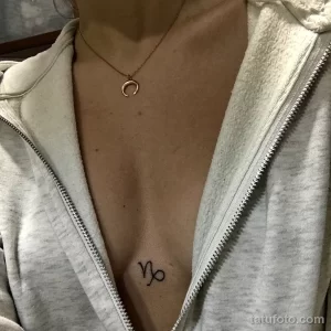 Фото тату: маленькая, черная татуировка символа Козерога, расположенная на груди под ключицей.
