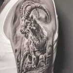 Черно-серая реалистичная татуировка на плече с изображением головы козерога, стоящего на скале, выполненная с использованием техники штриховки и растушевки.