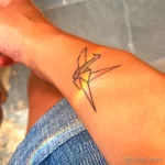 Фото минималистичной татуировки оригами журавля на запястье. Изображение выполнено тонкими черными линиями с добавлением желтой подсветки.