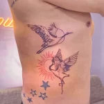 Фото тату с изображением двух журавлей в полете на фоне луны и звезд. Лаконичный дизайн в стиле line art, выполненный в черном цвете. Идеально для любителей минимализма.