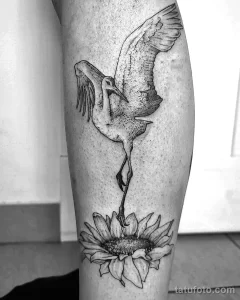 Фото тату на ноге с изображением аиста, стоящего на цветке лотоса. Черно-белый рисунок в графическом стиле, символизирующий чистоту и духовное просветление.