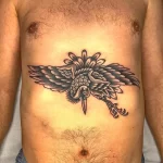 Фото тату на груди мужчины с изображением ворона с расправленными крыльями, окруженного цветами, выполненное в традиционном стиле.