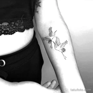 Фото тату с изображением двух минималистичных журавлей в полете, выполненных тонкими черными линиями на предплечье девушки.