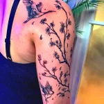 Фото тату с изображением летящего журавля и ветви цветущей сакуры, выполненное в технике лайнаворк на плече девушки.