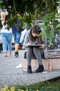 Фото тату: женщина, увлеченно рисует эскиз татуировки, сидя на скамейке в парке, окруженная зеленью.