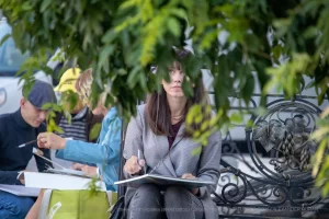 Фото тату: женщина-художник рисует эскиз татуировки на улице, сосредоточенно глядя в блокнот. На заднем плане другие люди и зелень.