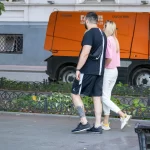 Фотография мужчины и женщины, идущих по улице, с видимой татуировкой на ноге мужчины. Яркий солнечный день, городская среда.