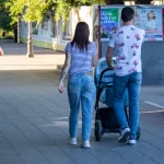Фото тату на руке женщины, идущей с мужем и ребенком в коляске по городской улице. Рисунок татуировки выполнен в черном цвете и частично виден из-под рукава.