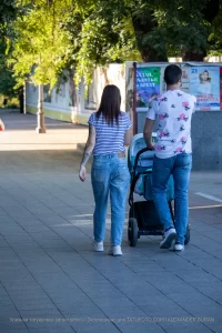 Фото тату на руке женщины, идущей с мужем и ребенком в коляске по городской улице. Рисунок татуировки выполнен в черном цвете и частично виден из-под рукава.