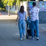 Фото тату на руке женщины, идущей с мужем и ребенком в коляске по городской улице. Рисунок татуировки выполнен в черном цвете и частично виден из-под рукава.