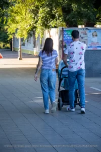 Фото тату на руке женщины, идущей с мужем и ребенком в коляске по городской улице. Рисунок татуировки выполнен в черном цвете и частично виден из-под рукава.