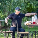 Фото мужчины с татуировками на руках и шее, сидящего за столиком в кафе. Мужчина одет в черную футболку и кепку.