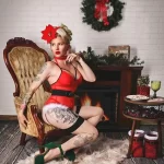 Фотография девушки в рождественском образе с видимыми татуировками на руке и ноге. Красивая и стильная модель позирует на фоне камина.
