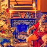 Фото мужчины в костюме Санты Клауса, сидящего у камина с татуировками на теле. Мужчина выглядит расслабленно и уверенно.