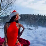 Фотография девушки в красном комбинезоне и шапке Санты, с множеством татуировок на руках и теле, на фоне заснеженного зимнего пейзажа.