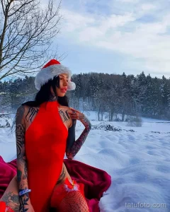 Фотография девушки в красном комбинезоне и шапке Санты, с множеством татуировок на руках и теле, на фоне заснеженного зимнего пейзажа.