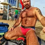 Фото мужчины в шапке Деда Мороза на пляже с татуировками на теле. Юмористический образ и летняя атмосфера.