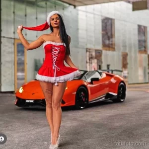 Сексуальная девушка в красном костюме Санты позирует рядом с роскошным оранжевым автомобилем, демонстрируя татуировку на бедрах.