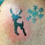 Фото минималистичной татуировки на плече с изображением оленя и снежинки, выполненной в синих тонах.