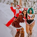 Фотография двух девушек и Санты в снежной обстановке, демонстрирующих татуировки на ногах и руках, создающие провокационный образ.
