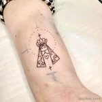 Фото татуировки с изображением Девы Марии в минималистичном стиле, с нимбом, крестом и точками, на руке.
