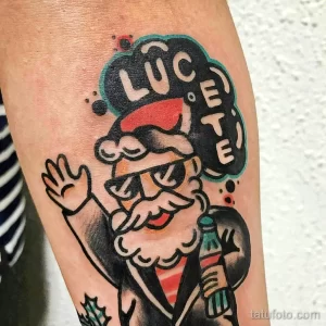 Фото тату Санты Клауса с надписью 'LUCETE' в стиле олдскул на руке мужчины. Яркие цвета и четкие контуры.