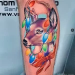 Яркая татуировка на предплечье с изображением головы оленя, украшенной разноцветными гирляндами.