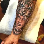 Фото тату: реалистичный портрет женщины, переходящий в изображение головы тигра на руке. Детализированный рисунок с яркими голубыми глазами.