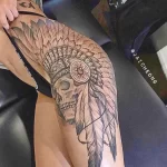 Фото тату на бедре: череп, украшенный перьями в стиле Native American. Детализированный черно-серый рисунок татуировки с акцентом на текстуру перьев.