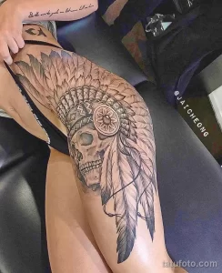 Фото тату на бедре: череп, украшенный перьями в стиле Native American. Детализированный черно-серый рисунок татуировки с акцентом на текстуру перьев.
