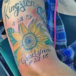 Фото яркой татуировки на руке с изображением подсолнуха и имен 'Kingston' и 'Graclynn' с датами. Татуировка выполнена в ярких цветах, создающих красочный рисунок.