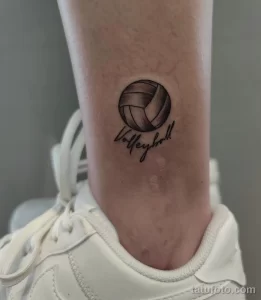 Фото тату: небольшой черный рисунок волейбольного мяча с надписью 'Volleyball' на лодыжке. Простой и стильный эскиз татуировки.