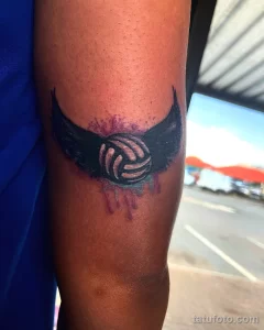 Фото татуировки волейбольного мяча с черными крыльями и фиолетовыми брызгами на руке. Эффект акварели.