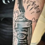 Фото тату бутылки Jack Daniel's с сердечками и надписями, выполненное в смешанном стиле. Оригинальный и эмоциональный рисунок татуировки на руке.