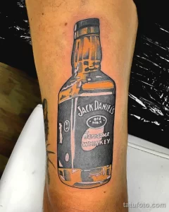 Черно-серая татуировка на руке с изображением бутылки Jack Daniel's. Детализированный рисунок татуировки с реалистичной прорисовкой этикетки.