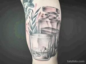 Черно-серая татуировка на предплечье с изображением бутылки и стакана, украшенных ветками. Детализированный рисунок татуировки в стиле реализм.