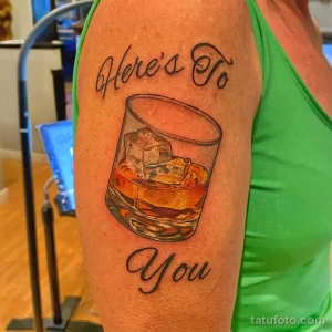 Фото тату стакана виски с кубиками льда и надписью 'Here's To You' на бицепсе. Реалистичный рисунок татуировки в коричнево-черных тонах.