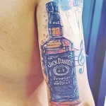 Фото татуировки на руке с реалистичным изображением бутылки виски Jack Daniel's, выполненной в ярких цветах.