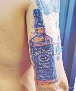 Фото татуировки на руке с реалистичным изображением бутылки виски Jack Daniel's, выполненной в ярких цветах.
