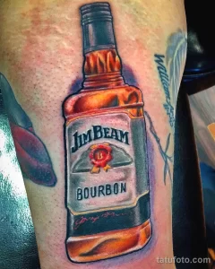 Фото тату реалистичной бутылки виски Jim Beam на мускулистом предплечье. Детализированный рисунок с яркими оранжевыми и коричневыми оттенками.