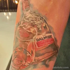 Фото татуировки бутылки виски The Dalmore с изображением оленьего рога. Реалистичный стиль с использованием черно-серых тонов.