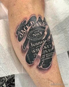 Фото реалистичной татуировки бутылки виски Jack Daniel's на руке. Детализированное изображение с тенями и текстурами.