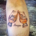 Фото тату на ноге с изображением двух бокалов с виски и надписью 'George'. Минималистичный стиль и коричнево-белая цветовая гамма.