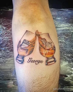 Фото тату на ноге с изображением двух бокалов с виски и надписью 'George'. Минималистичный стиль и коричнево-белая цветовая гамма.
